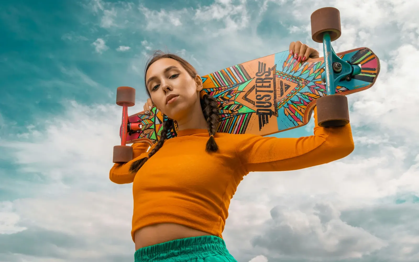 Skater holding a patterned longboard across her shoulders under a bright sky